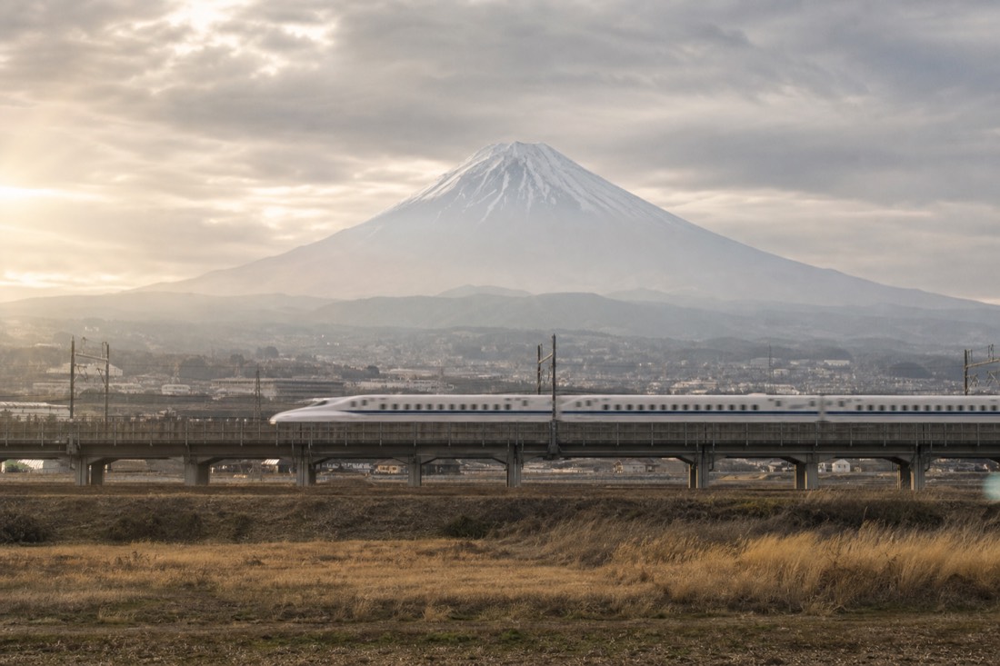 A high-speed train crossing farmland with the pale cone of Mount Fuji rising in the haze behind it
