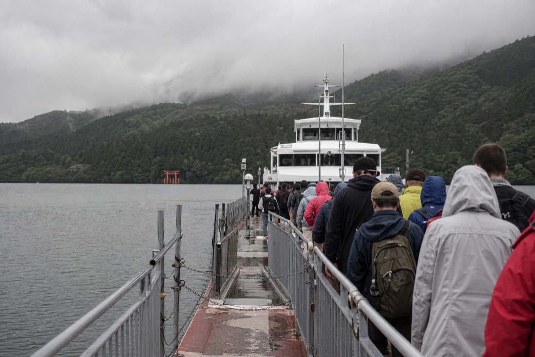 A queue of visitors in raincoats boarding a large white sightseeing boat at a grey lakeside pier