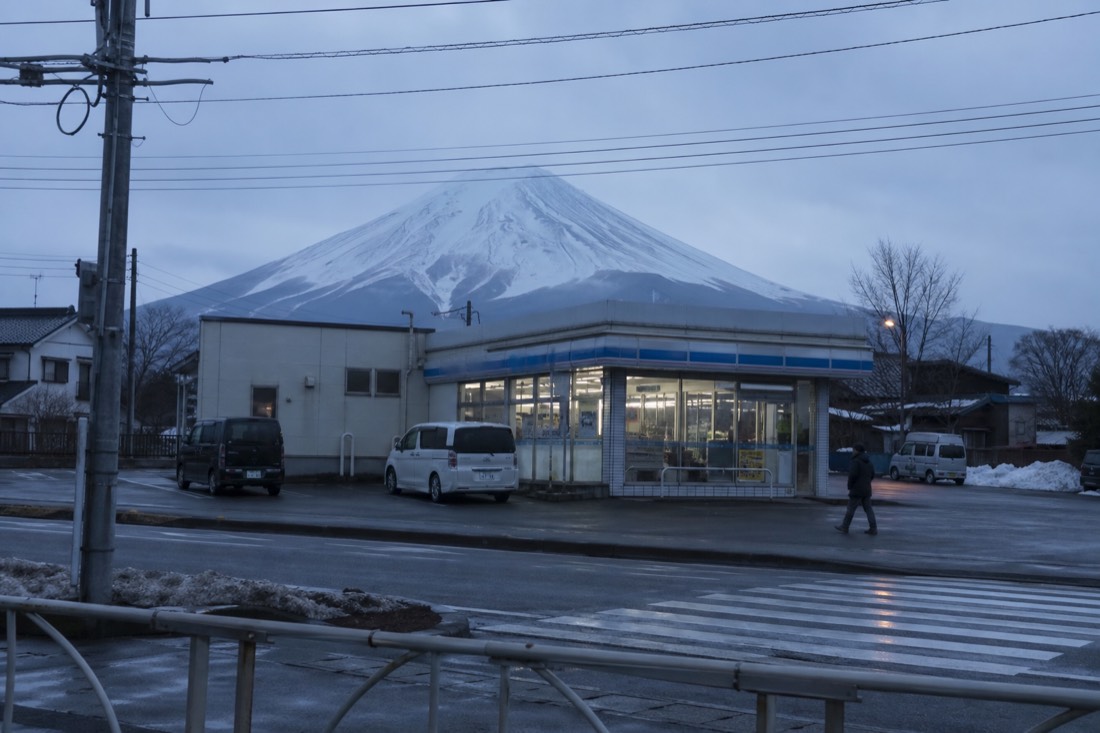 A low convenience store on a small-town street with the snow-capped cone of Mount Fuji rising directly behind its roof